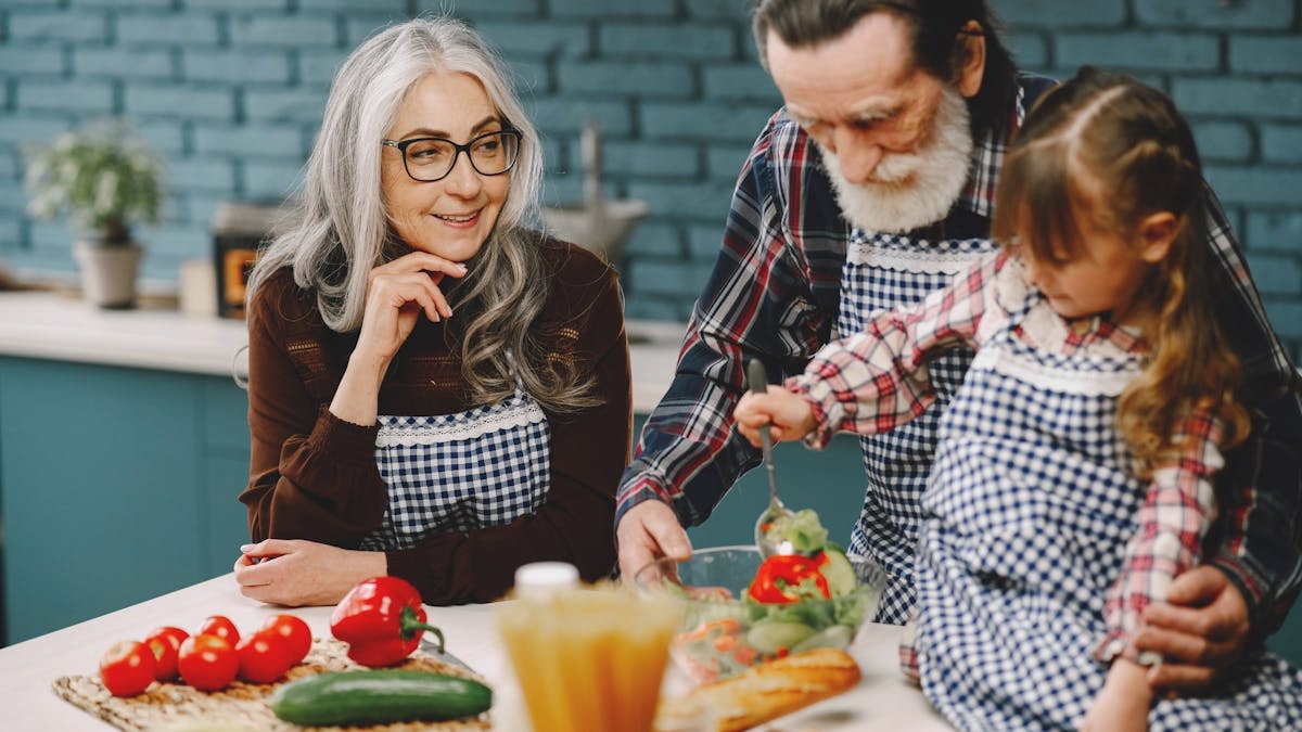 Grandparents and a child enjoy cooking together in a warm kitchen setting.